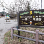 There is a sign advertising Hida Beer at a brewery, in Japan situated by a peaceful country road lined with old fashioned houses and trees in the backdrop.