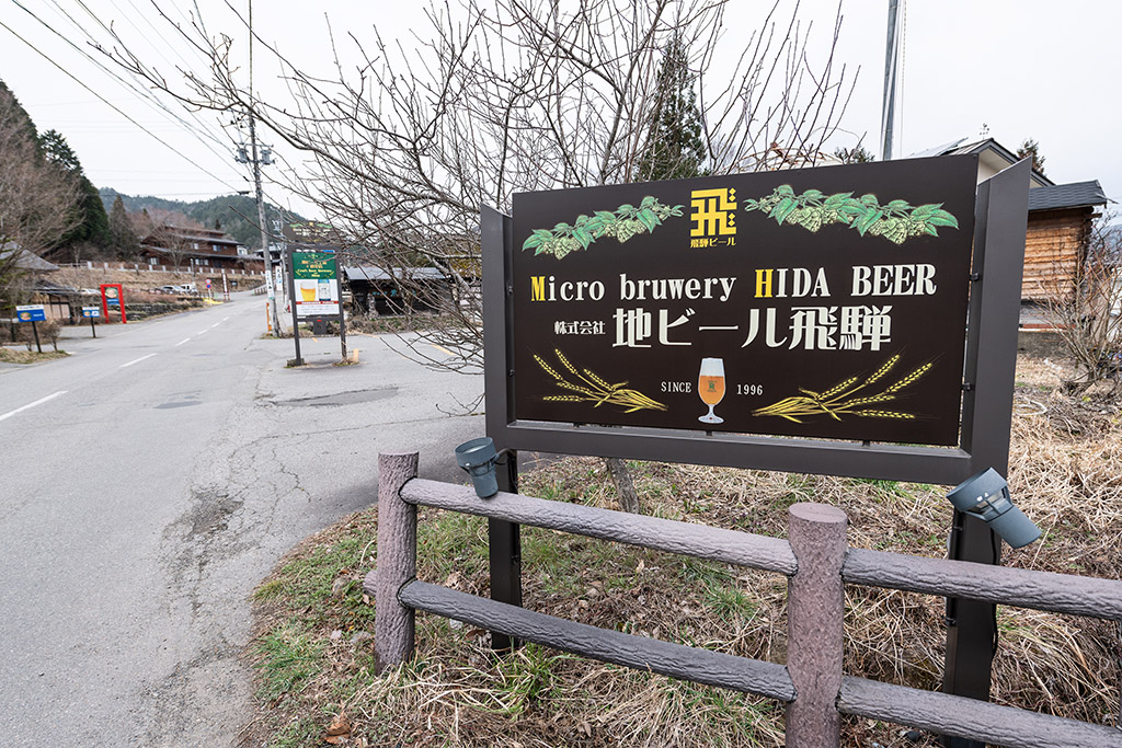 There is a sign advertising Hida Beer at a brewery, in Japan situated by a peaceful country road lined with old fashioned houses and trees in the backdrop.